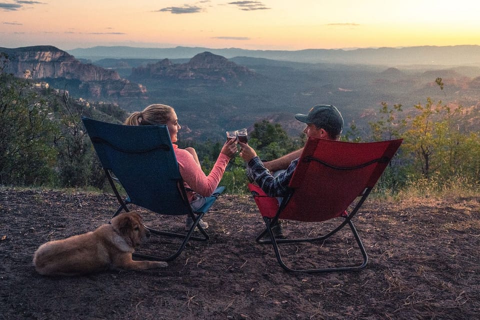 Man and woman sitting on red and blue camping chairs, each with a glass of wine in front of a rugged mountain landscape and a yellow dog at their feet.