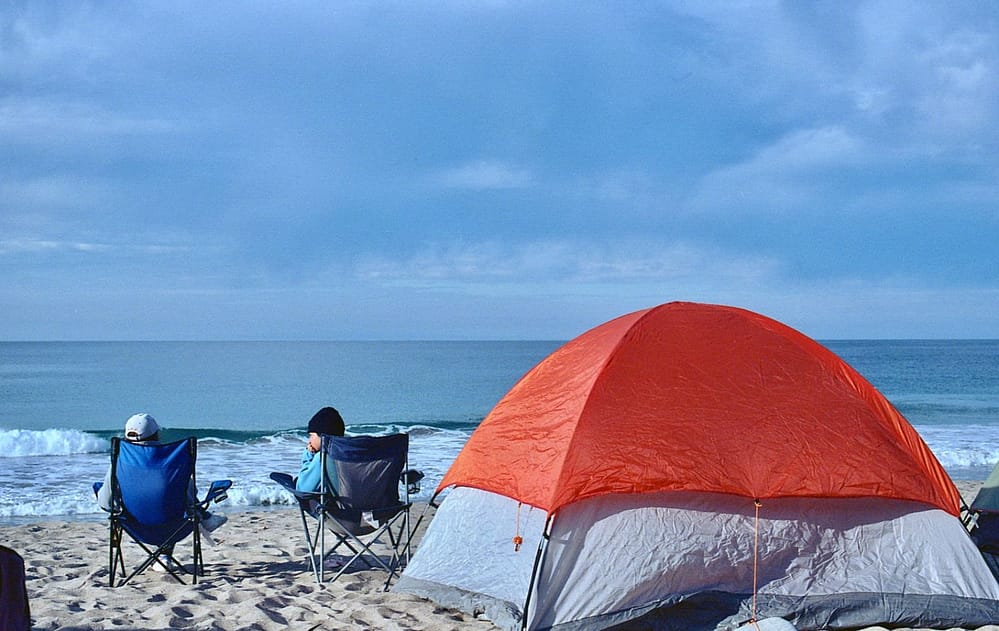 Man and woman sitting on camping chair on beach facing the ocean with a red camping tent behind them
