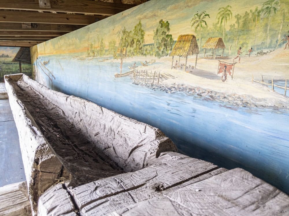 Replica of a dugout canoe with a painted background of Seminole village
