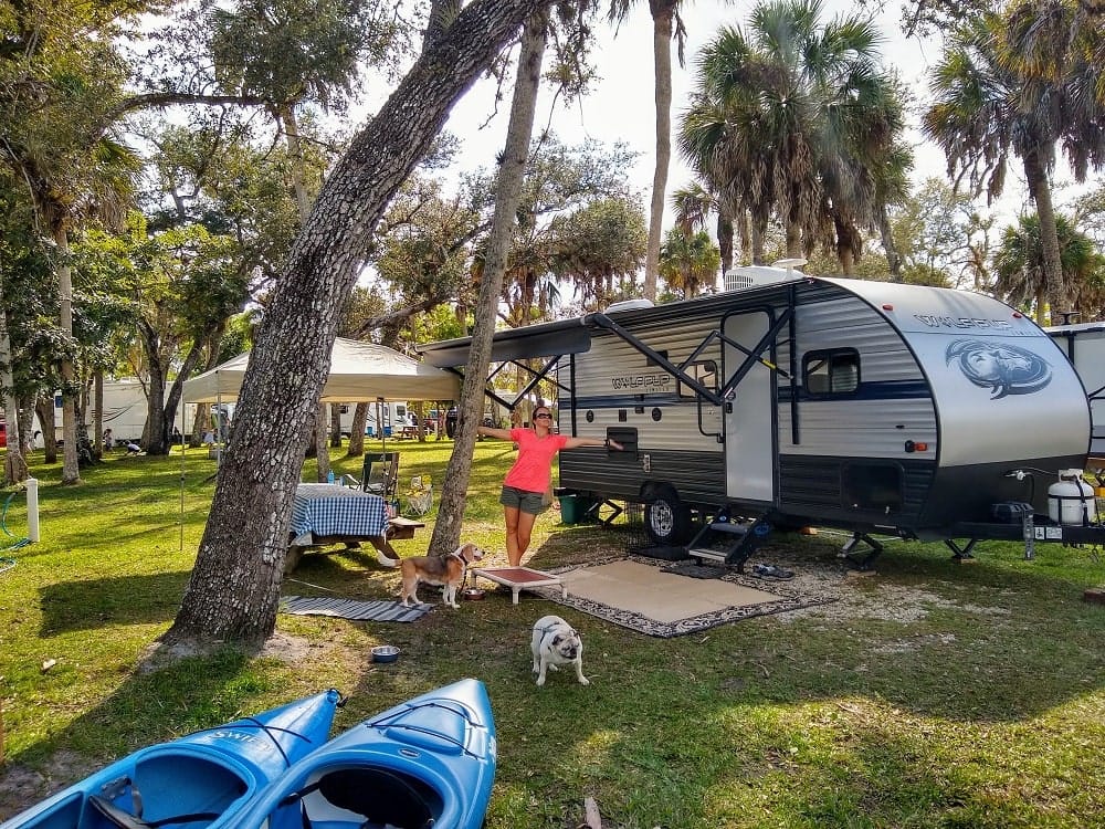 Woman in front of travel trailer, two blue kayaks and two small dogs