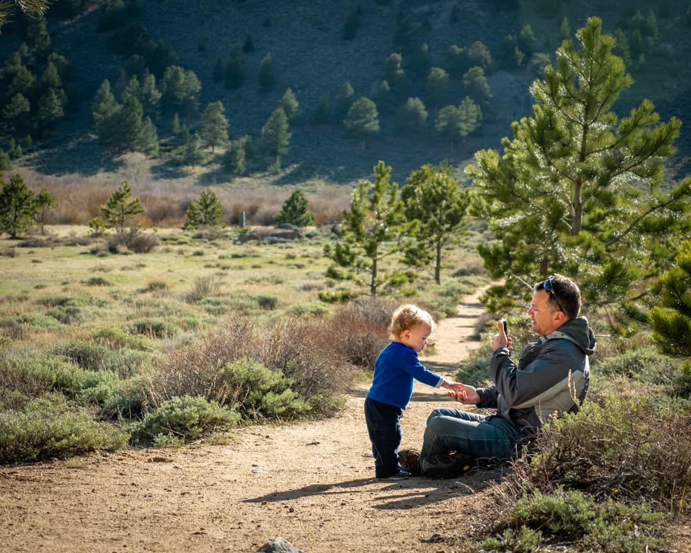 man sitting down taking photo of toddler on a trail
