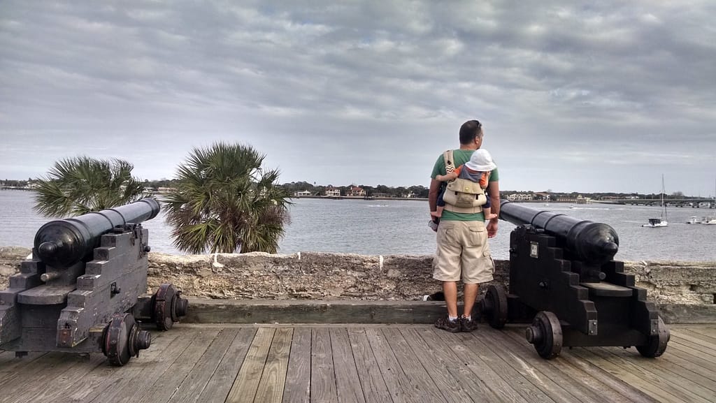 Man carrying infant on baby carrier on his back while standing between two cannons in Castillo de San Marco, St. Augustine, FL.