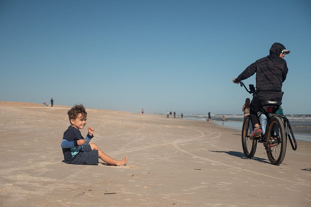 Small child sitting and smiling on sand as a person on a bicycle rides past 