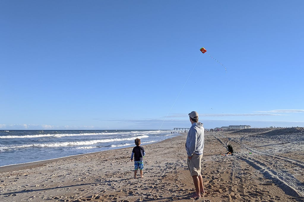 man wearing a light gray hoodie observing a small child flying a small colorful kite on a large expanse of sand next to the ocean under clean blue sky