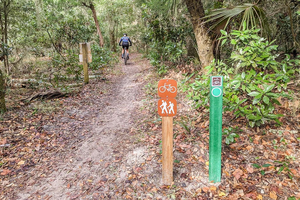 Beginning of a nature trail with a sign showing hikers and bike. In the background a person riding a bike on the trail