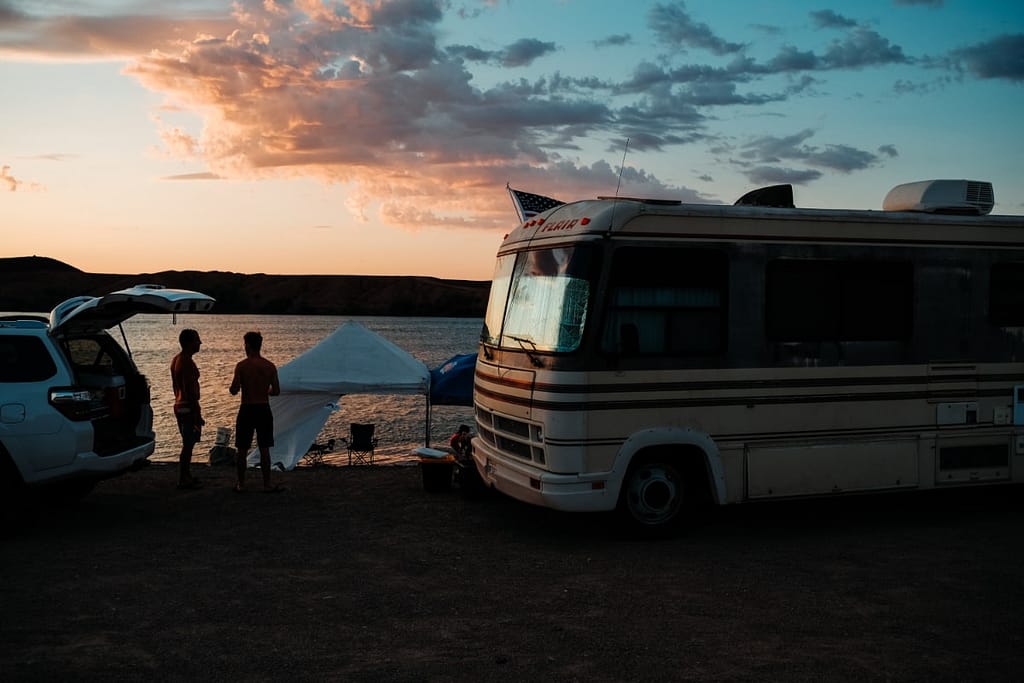 Silouhettes of two people next to a motorhome, with a screenhouse in the background against sunset skies