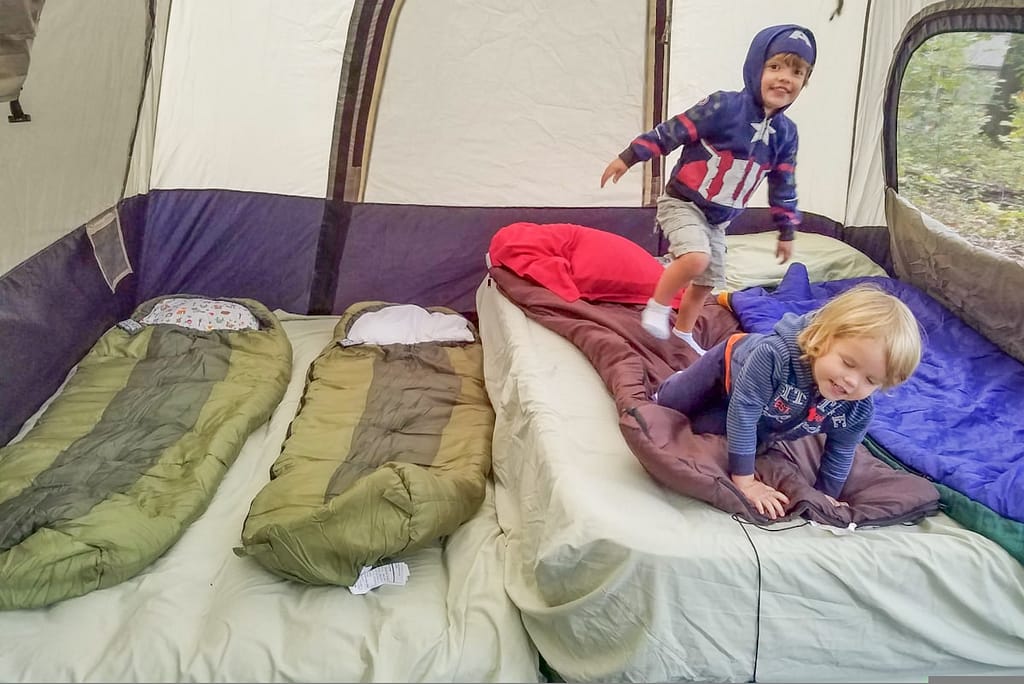 A boy and a toddler girl playing on a large inflatable mattress with sleeping bags inside a large camping tent